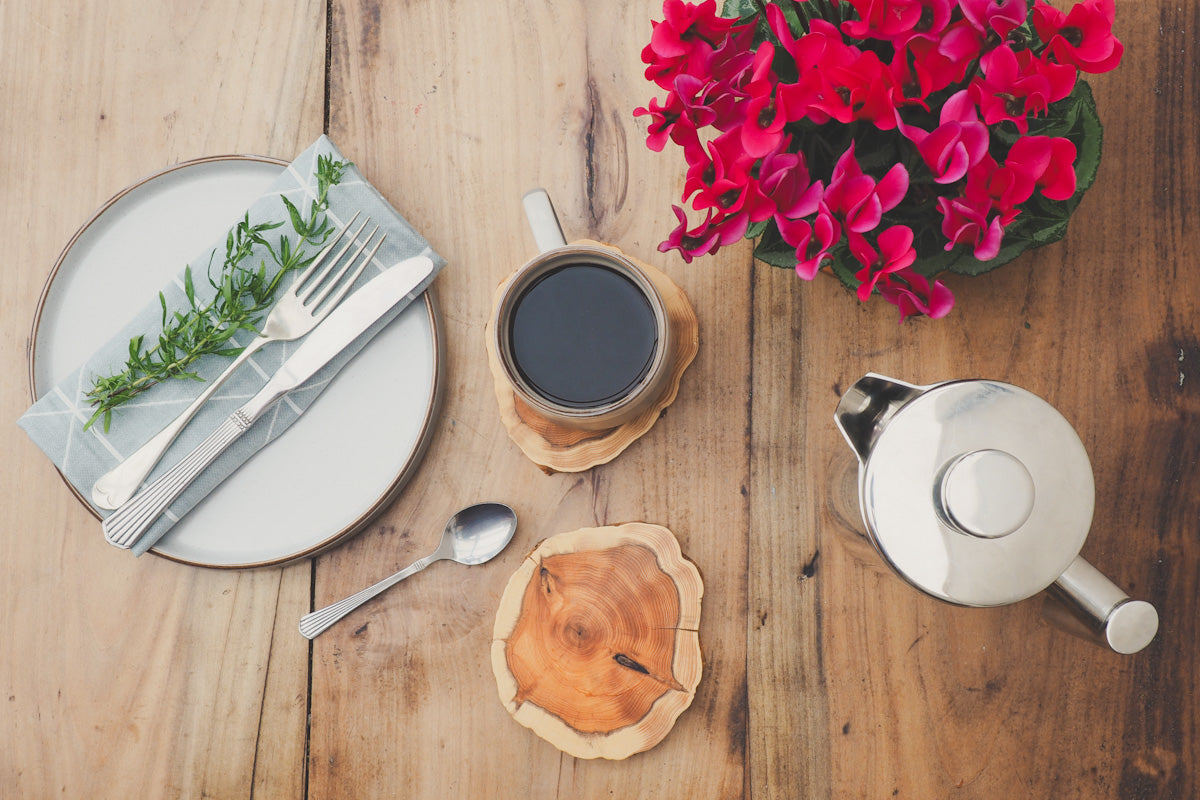 Looking down on a table with plates, flowers, cafetière and a mug of coffee on a wooden yew coaster 